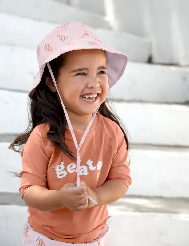 Ein lachendes Mädchen mit dunklen Haaren steht auf einer Treppe. Es trägt den UV-Sonnenhut Ice Cream von Lässig und dazupassenden Badehosen
