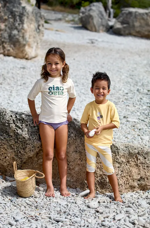 Zwei Kinder am Kieselstrand. Das grössere Mädchen trägt ein UVSchutz Shirt von Lässig in offwhite mit grünem Schriftzug "Ciao"