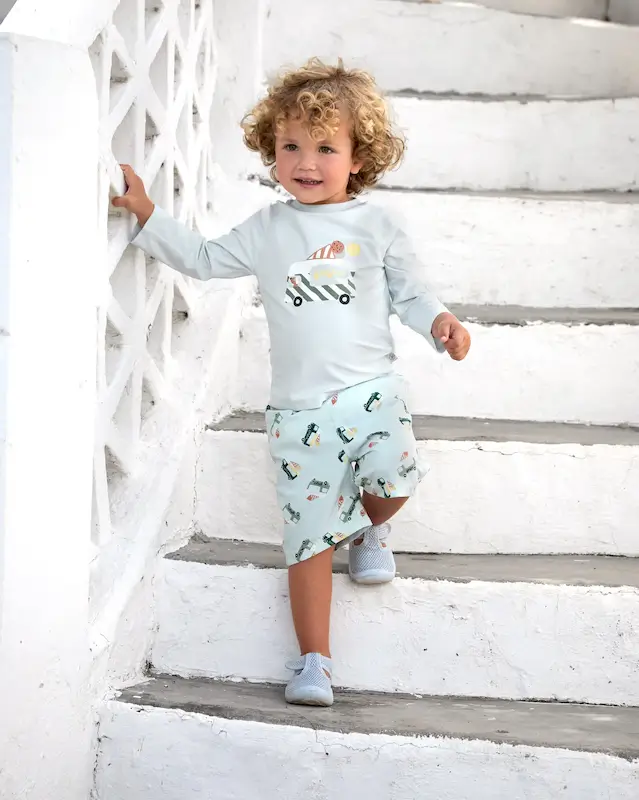 Ein kleiner Junge steigt eine Treppe herunter. Er trägt hellblauer UV-Badehosen und ein Sonnenschutzshirt  mit bunten Eiswagen von Lässig.