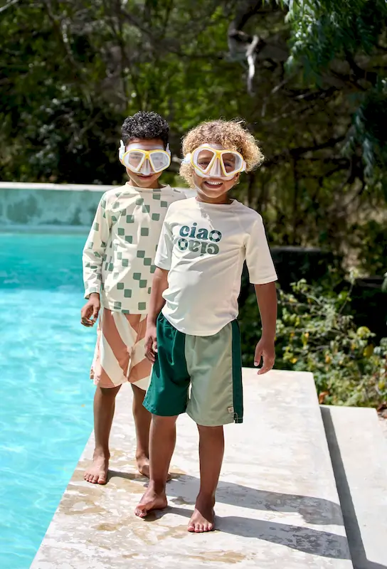 Zwei Jungen am Pool, der vordere Junge trägt das weisse UV-Shirt "Ciao" und grüne Badehosen von Lässig.