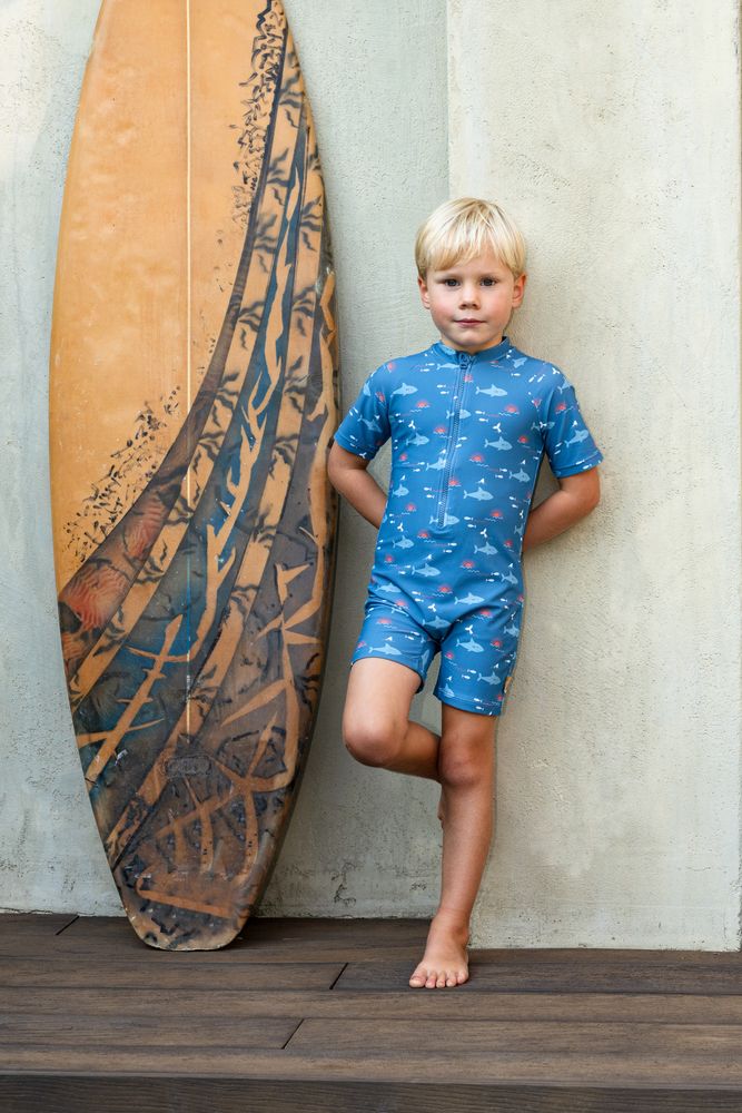 Ein Junge lehnt neben einem gelben Surfbrett an einer Wand. Er trägt den UV Swimsuit "Shark" von Fresk