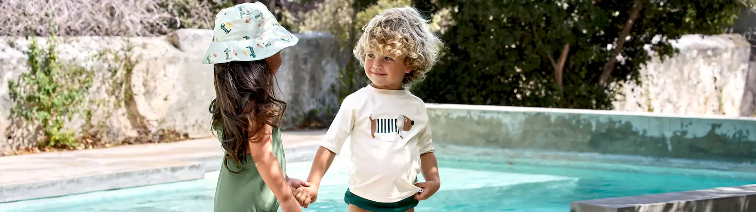 Zwei Kinder in Lässig UV-Badeshirts stehen Hand in Hand am Pool.