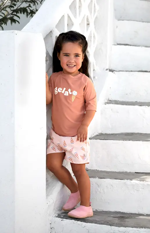 Ein Mädchen steht am Fuss einer weissen Aussentreppe. Sie trägt die Lässig Mädchenbadeshorts Ice Cream Pink und ein UV-Shirt von Lässig