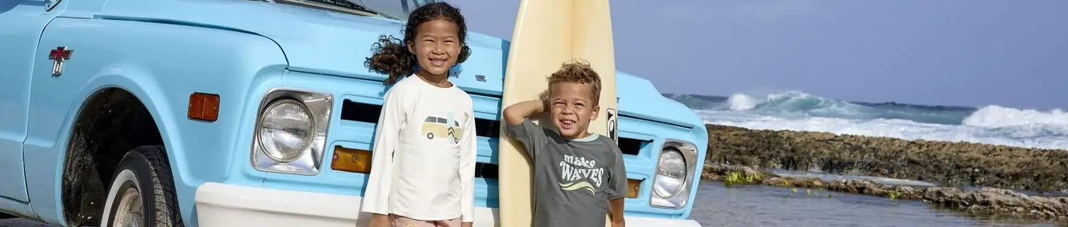 Zwei Kinder in Lässig Badekleidern vor einem hellblauen Auto am Strand.