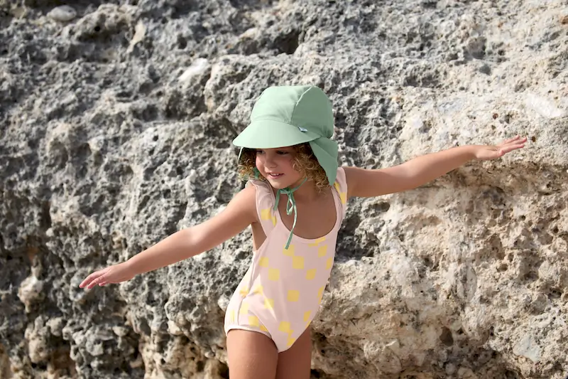 Mädchen mit Sonnenhut Green und Badkleid von Lässig steht an einem Felsen am Strand