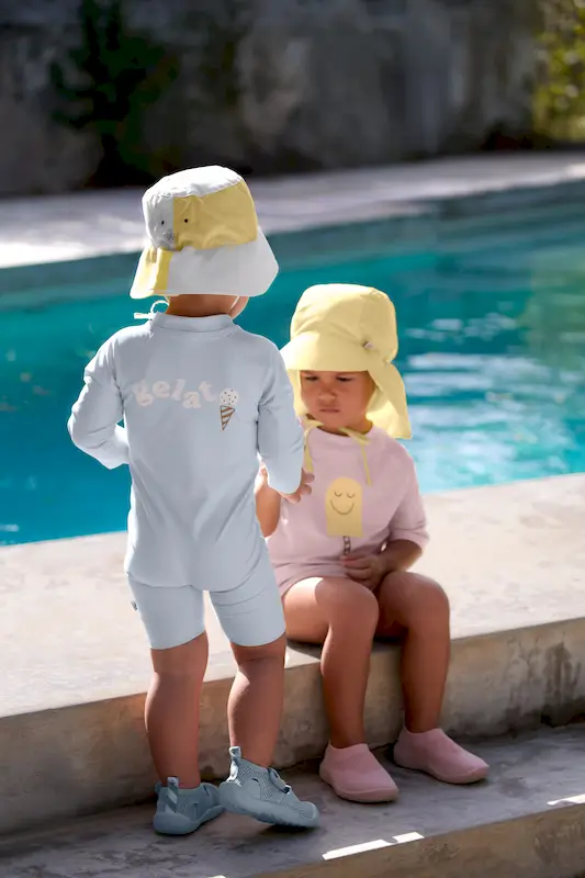 Zwei Kinder sind an einem Pool. Eines trägt die hellgelbe UV-Schirmmütze  Pale Yellow mit Nackenschutz von Lässig.