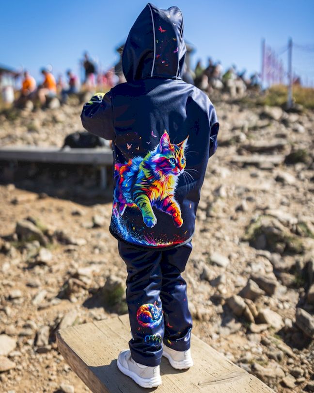 Kind steht auf einem Holzbrett im Freien und trägt dunkelblaue Softshellhose mit passender Jacke mit buntem Katzenmotiv auf der Rückseite