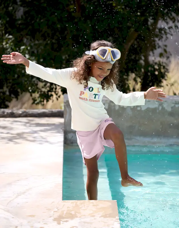 Mädchen am Pool planscht im Wasser, sie trägt ein UV-Shirt Langarm "Pool Party Sea Salt" und Badehosen von Lässig.