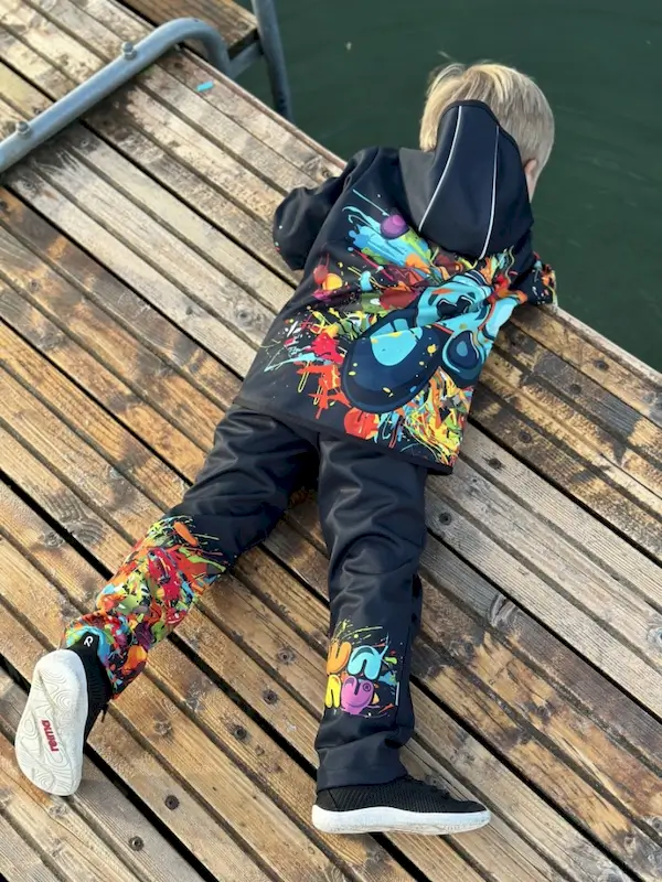 Ein Kind liegt auf einem Holzsteg auf dem Buch und blickt ins Wasser. Es trägt die Wamu Regenhose "Game On" in schwarz mit buntem Gamingmotiv