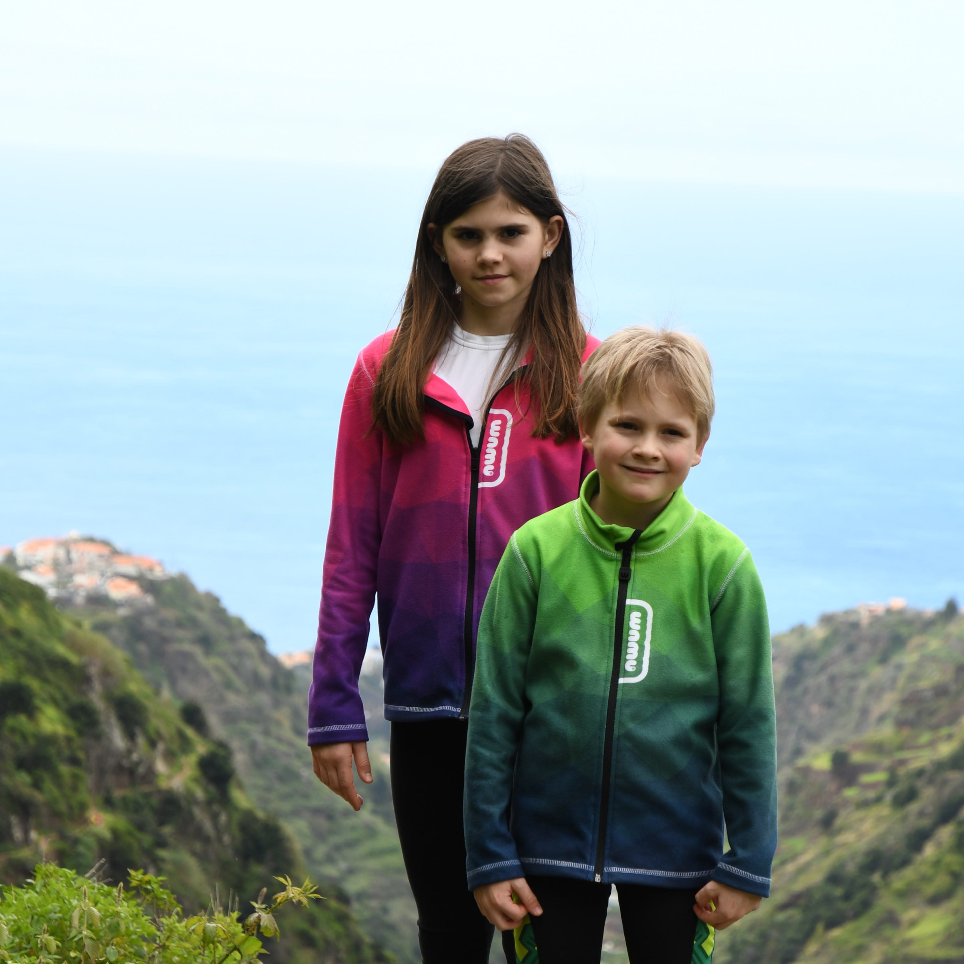 Mädchen und Junge auf einem Berg, im Hintergrund das Meer. Sie tragen die Fleecejacken Mosaic Purple und Mosaic Green.