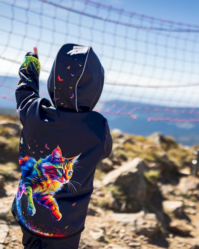 Mädchen mit dunkelblauer Softshelljacke mit buntem Katzenmotiv an einem steinigen Strand, vor einem hängenden Fischernetz