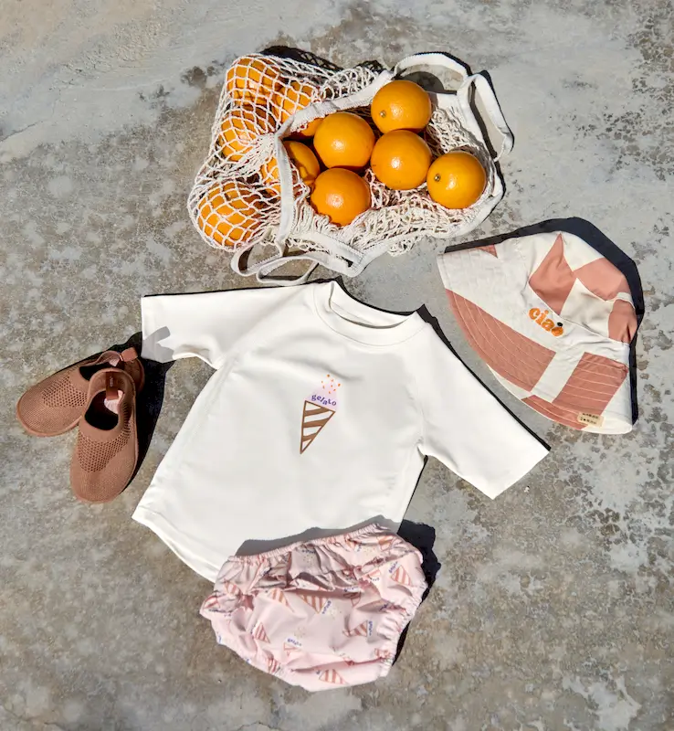 Flatlay der Lässig UV-Bademode "Ice Cream" mit weissem UV-Shirt Ice Cream mit Glacecornet, Schwimmwindeln, Sonnenhut, Badeschuhe und einem Netz Orangen.